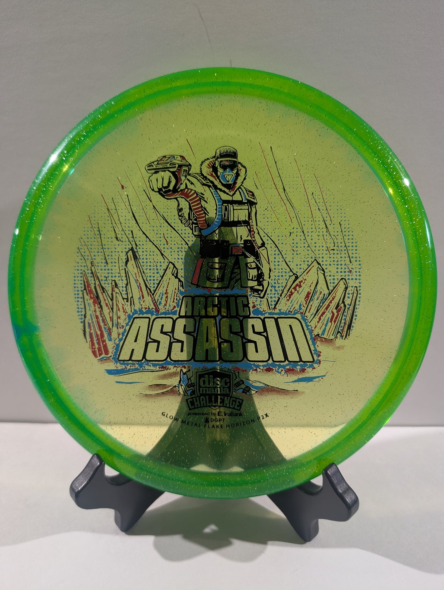 A Discmania Arctic Assassin disc golf disc with a green rim and a yellow center featuring a graphic of an Arctic soldier and mountains, displayed on a small stand.