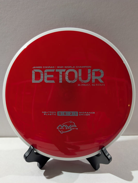Red disc golf driver with 'Detour' branding on a stand against a gray background