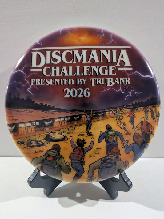 A disc for the Discmania Challenge 2026, presented by TruBank, displayed on a black stand against a neutral light gray background. The disc features a detailed illustration of people at a disc event under a stormy sky, with bold white text at the top.