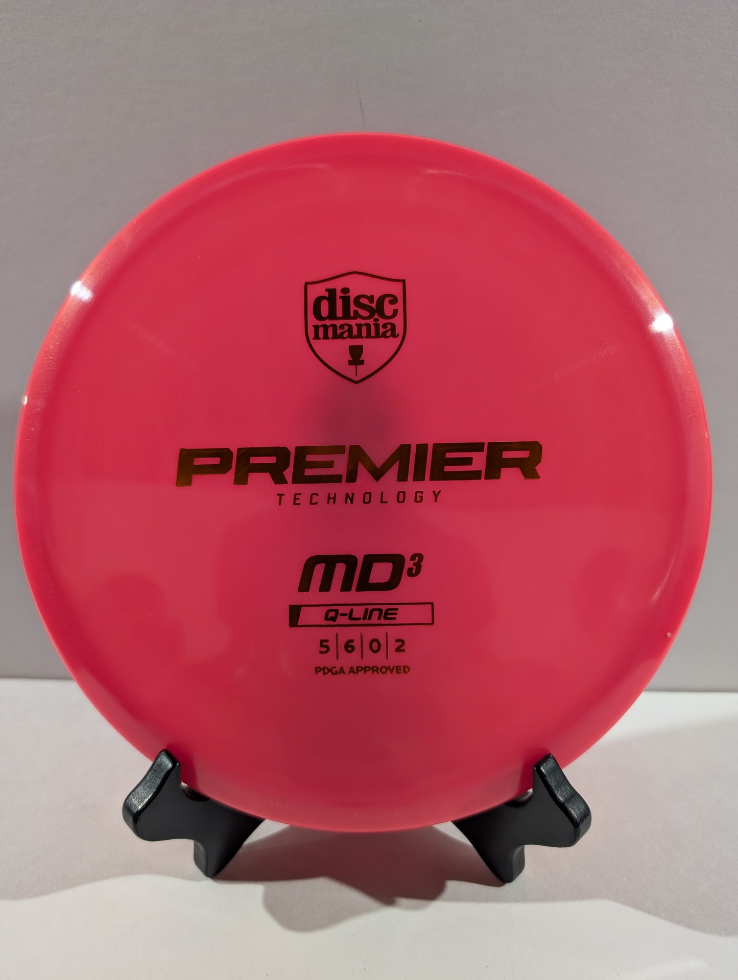 A bright pink Discmania Premier Technology MD3 Q-Line disc golf disc displayed on a small black stand against a neutral gray background.