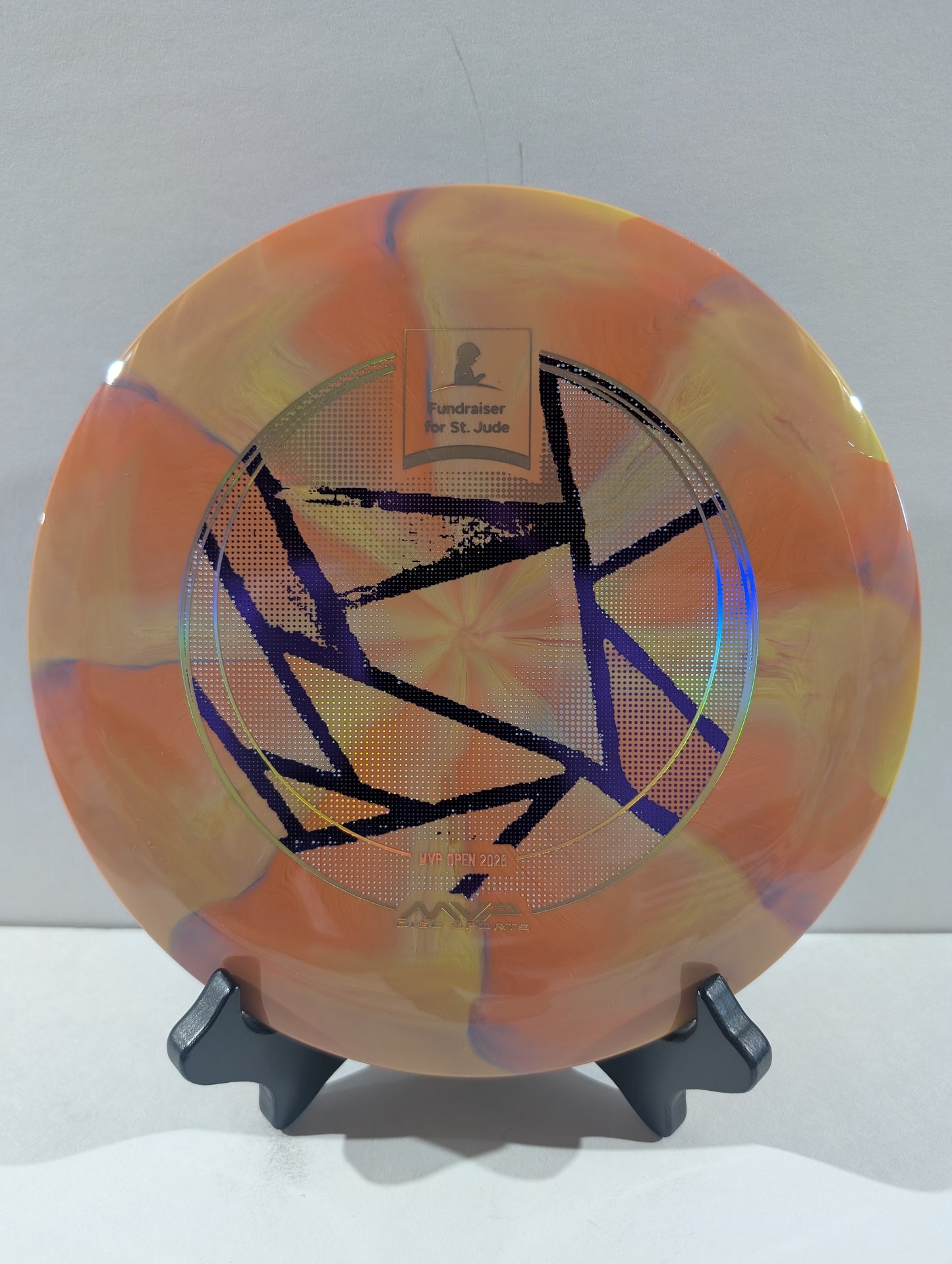 An orange disc golf disc with a marbled pattern of yellow, purple, and white, featuring a black geometric star design and a foil stamp in the center, displayed on a black stand against a light gray background.
