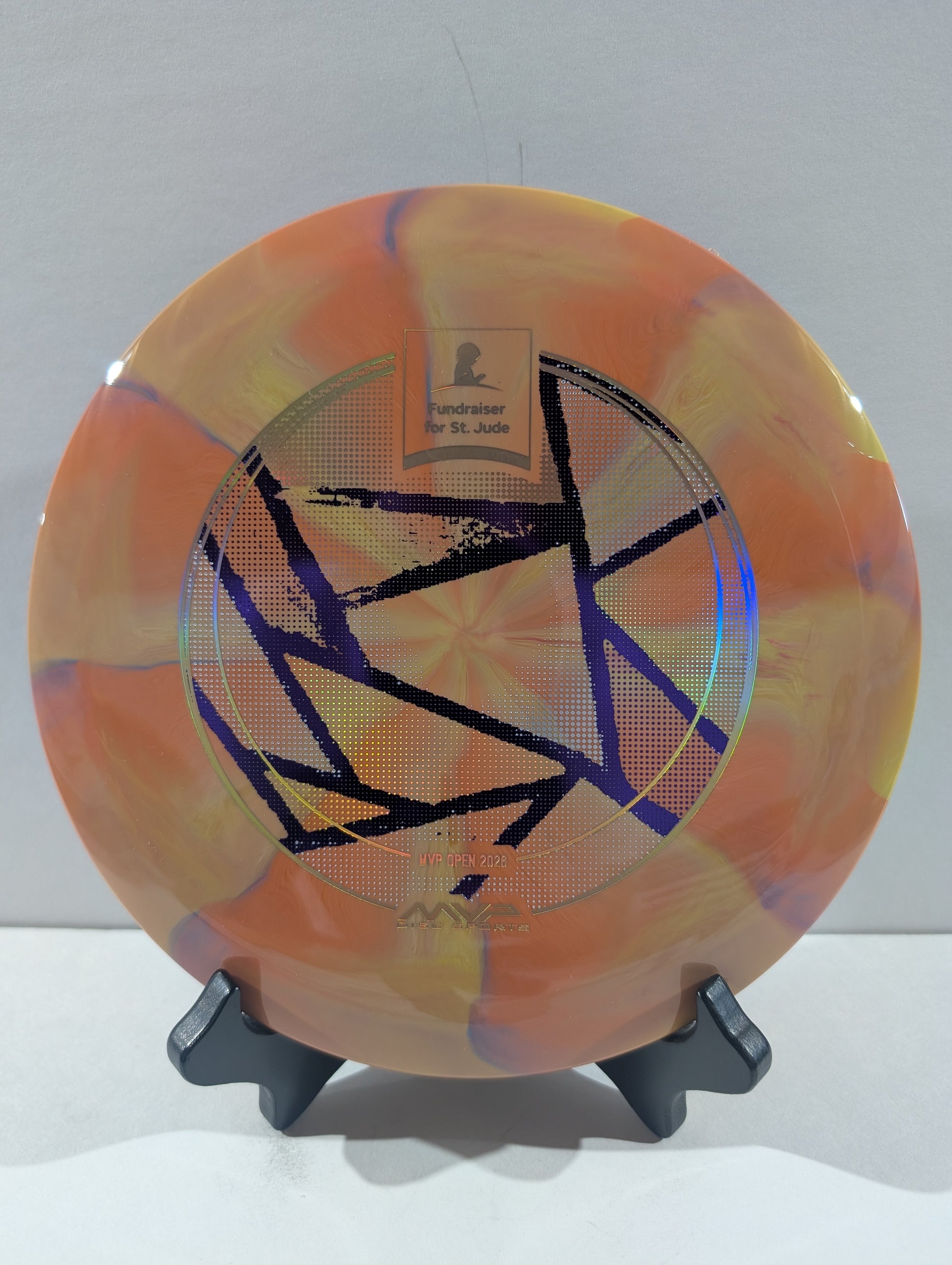 An orange disc golf disc with a marbled pattern of yellow, purple, and white, featuring a black geometric star design and a foil stamp in the center, displayed on a black stand against a light gray background.