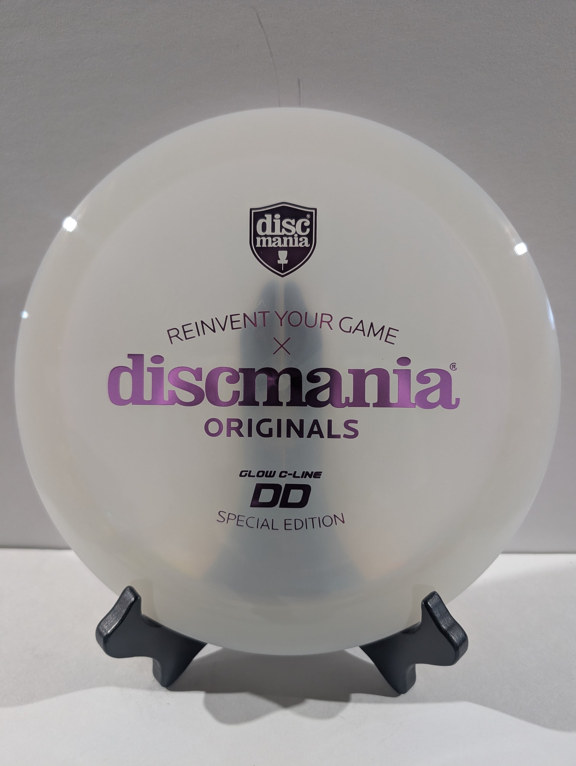 A white Discmania Originals DD Glow-C-Line Special Edition disc golf disc displayed on a black stand against a light gray background.