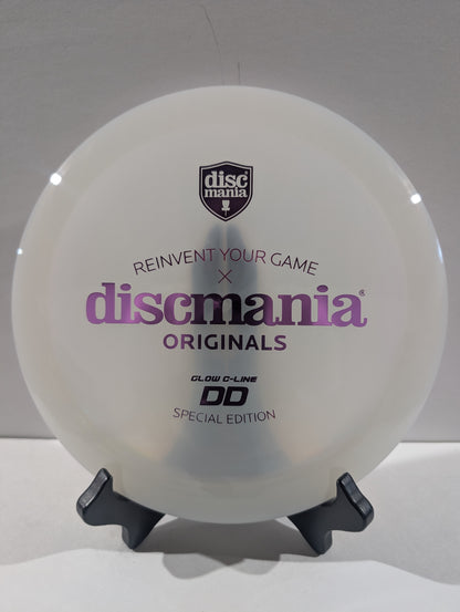 A white Discmania Originals DD Glow-C-Line Special Edition disc golf disc displayed on a black stand against a light gray background.