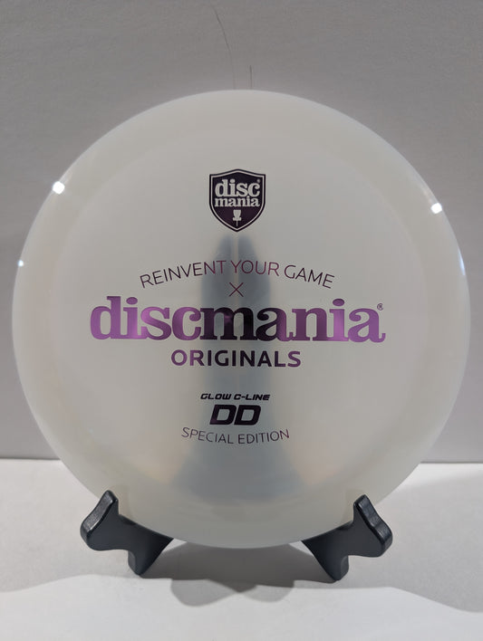 A white Discmania Originals DD Glow-C-Line Special Edition disc golf disc displayed on a black stand against a light gray background.