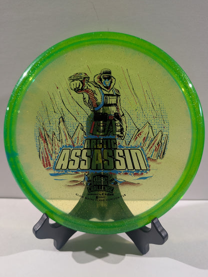 A Discmania Arctic Assassin disc golf disc with a green rim and a yellow center featuring a graphic of an Arctic soldier and mountains, displayed on a small stand.