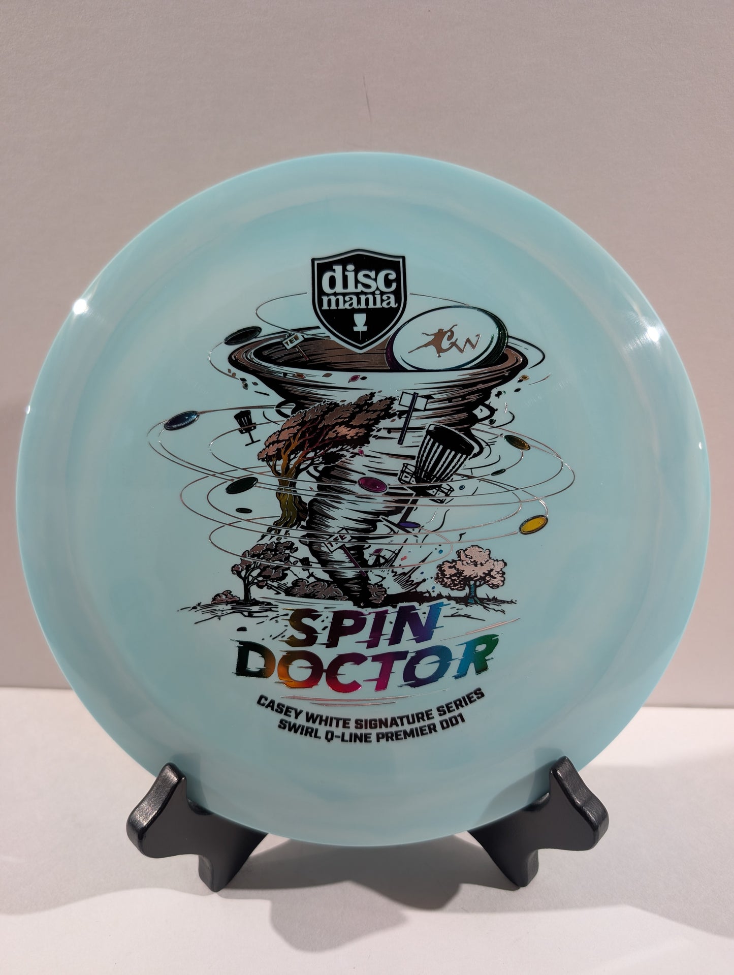 A light blue Discmania 'Spin Doctor' disc golf disc displayed on a black stand against a neutral beige background.