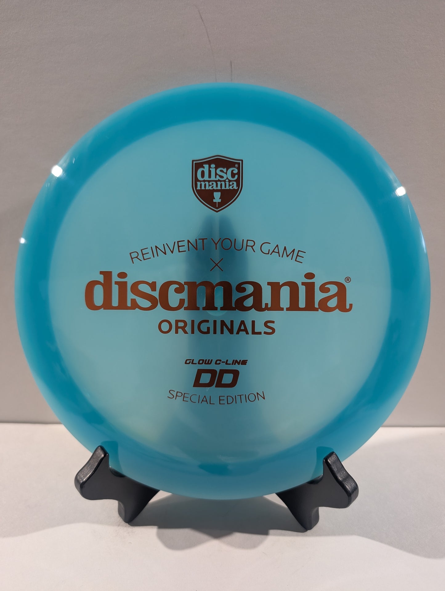 A Discmania Originals DD glow-in-the-dark special edition disc golf disc in a translucent light blue color, displayed on a small black stand against a neutral background.