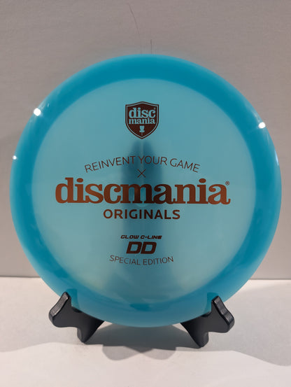 A Discmania Originals DD glow-in-the-dark special edition disc golf disc in a translucent light blue color, displayed on a small black stand against a neutral background.