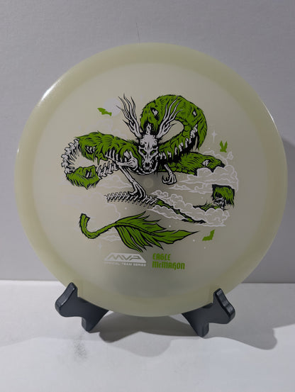 An MVP Disc Sports Eagle McMahon disc golf disc in translucent cream with a green dragon graphic, displayed on a black stand against a neutral gray background.