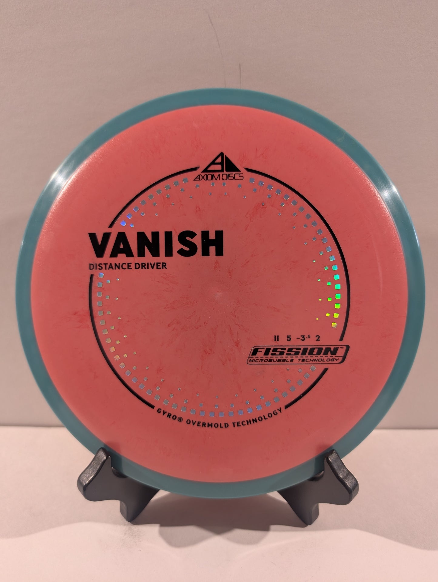 A disc golf disc, specifically a Fission Vanish distance driver, displayed on a small stand against a plain beige background. The disc features a coral-pink flight plate with a teal-blue outer rim and black printed text and graphics.
