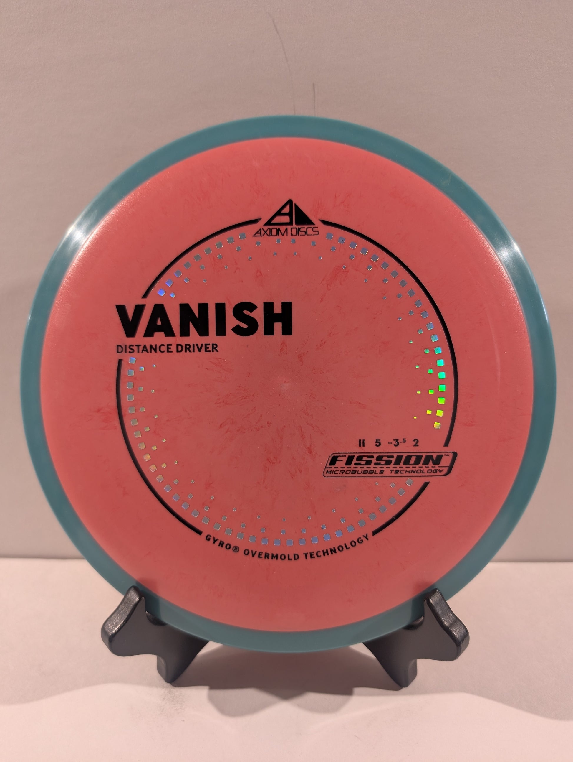 A disc golf disc, specifically a Fission Vanish distance driver, displayed on a small stand against a plain beige background. The disc features a coral-pink flight plate with a teal-blue outer rim and black printed text and graphics.