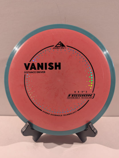 A disc golf disc, specifically a Fission Vanish distance driver, displayed on a small stand against a plain beige background. The disc features a coral-pink flight plate with a teal-blue outer rim and black printed text and graphics.