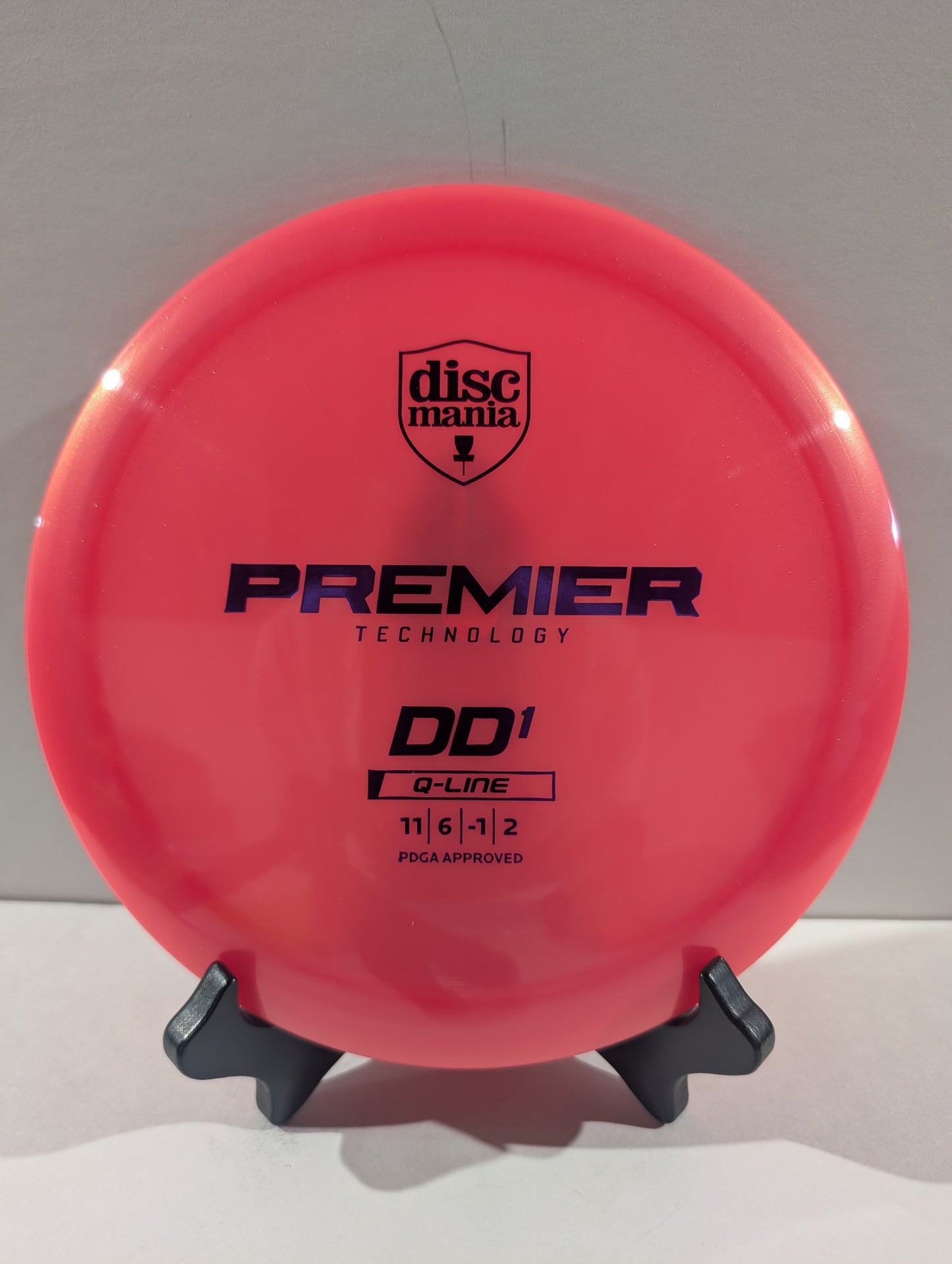 A bright pink Discmania Premier Technology DD1 Q-Line disc golf disc displayed on a small black stand against a light gray background.