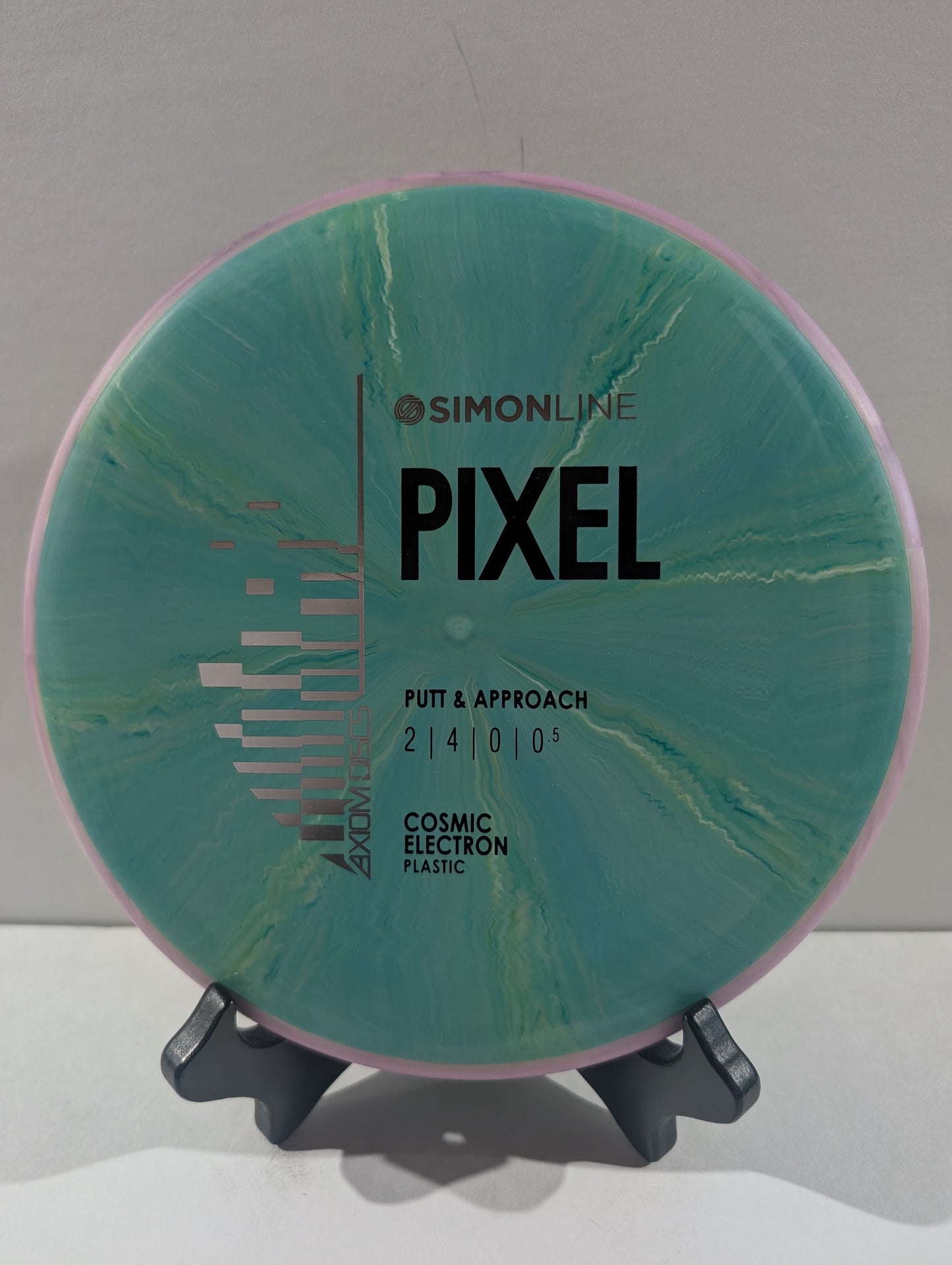 A disc golf putter and approach disc by Simon line Pixel, featuring a teal and green cosmic electron plastic design with a purple outer rim, displayed on a small stand.