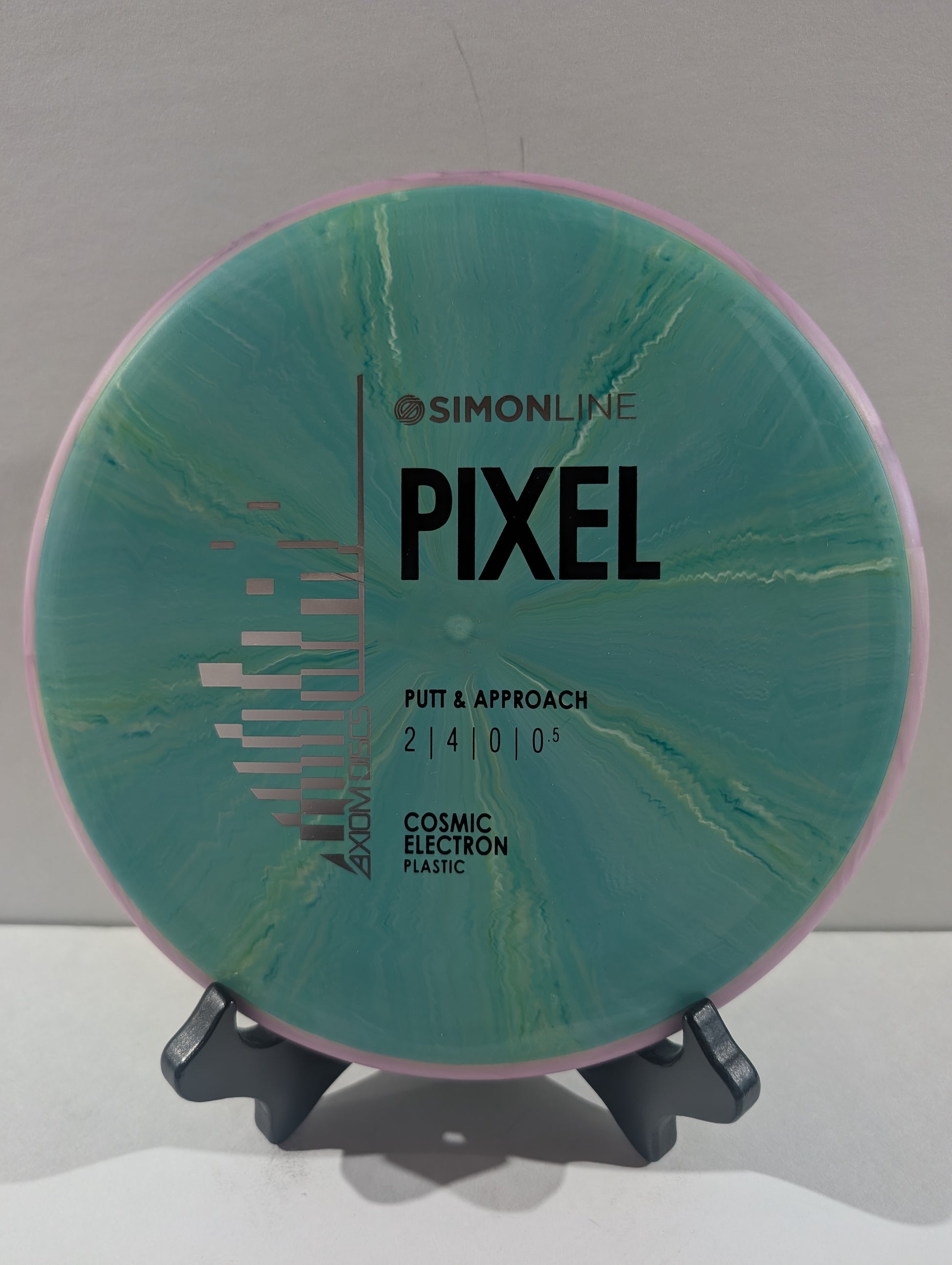 A disc golf putter and approach disc by Simon line Pixel, featuring a teal and green cosmic electron plastic design with a purple outer rim, displayed on a small stand.
