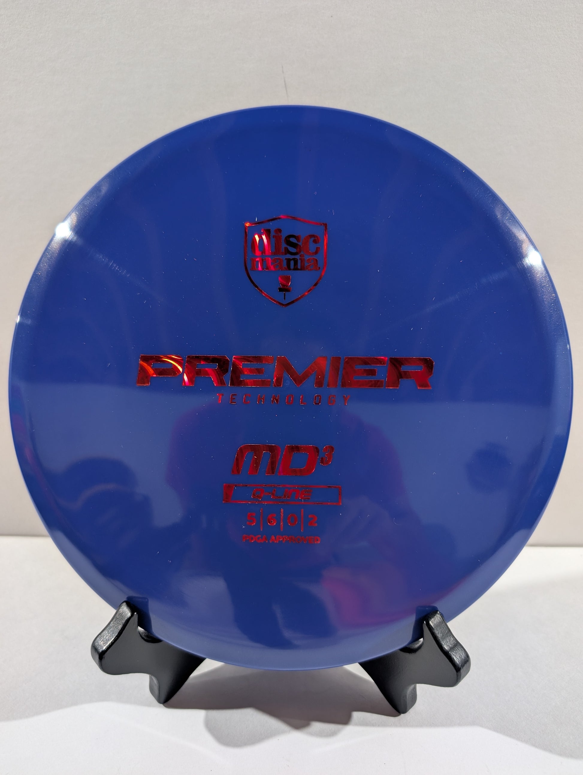 A blue Discmania Premier Technology MD3 Golf Disc with red foil stamping, displayed on a small black stand against a neutral light gray background.