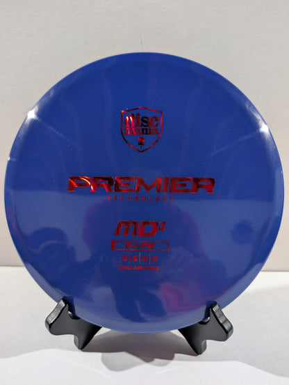A blue Discmania Premier Technology MD3 Golf Disc with red foil stamping, displayed on a small black stand against a neutral light gray background.