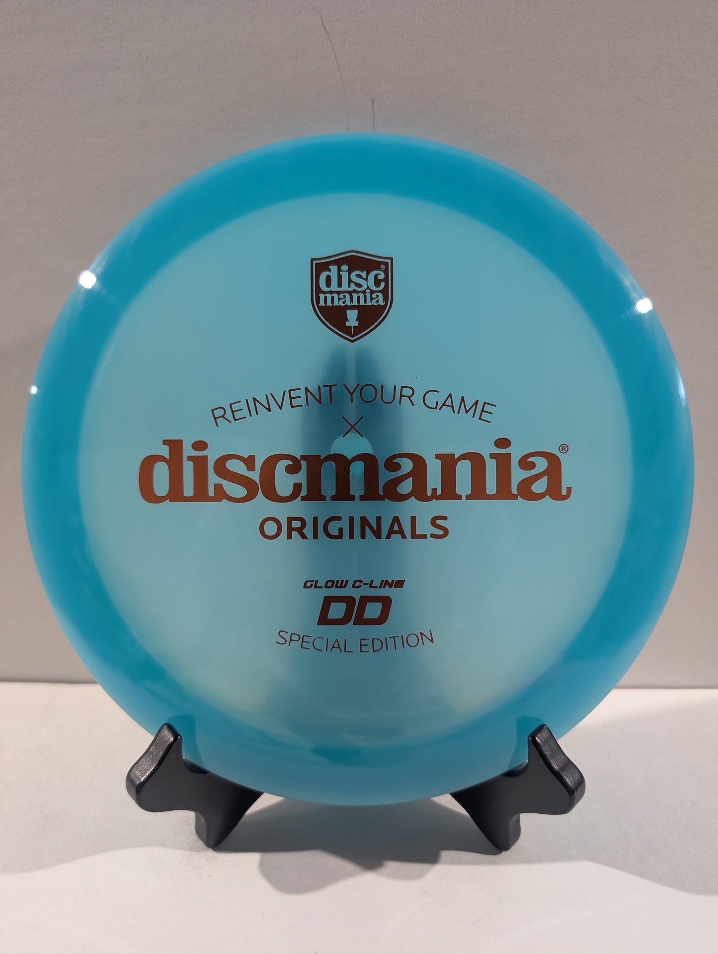 A translucent light blue Discmania Originals DD Glow C-Line disc golf disc displayed on a small black stand against a neutral gray background.