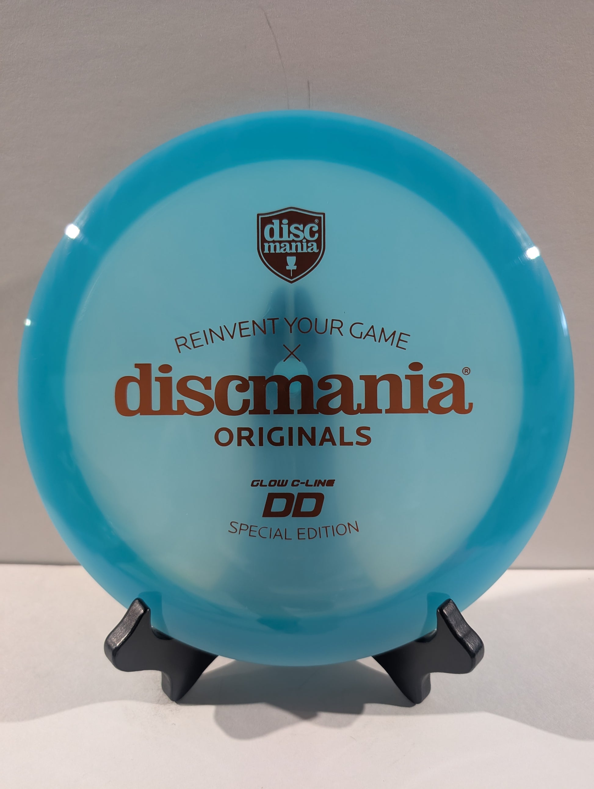 A translucent light blue Discmania Originals DD Glow C-Line disc golf disc displayed on a small black stand against a neutral gray background.