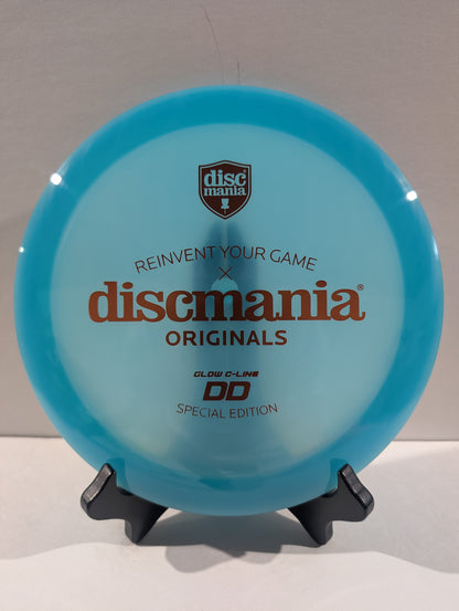 A translucent light blue Discmania Originals DD Glow C-Line disc golf disc displayed on a small black stand against a neutral gray background.