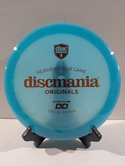 A translucent light blue Discmania Originals DD Glow C-Line disc golf disc displayed on a small black stand against a neutral gray background.