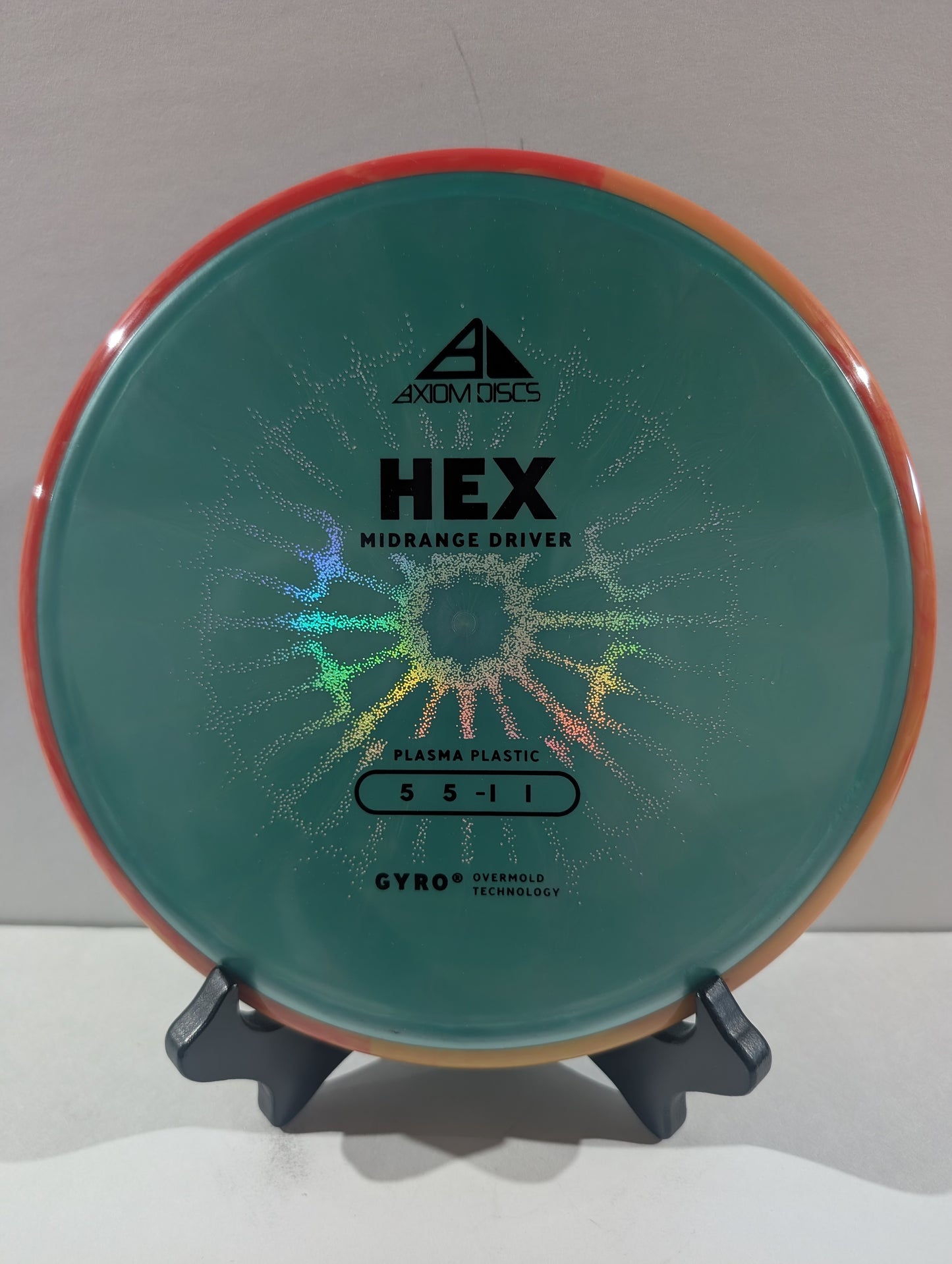 An Axiom Discs Hex Midrange Driver disc golf disc made of Plasma Plastic, featuring a turquoise center with a holographic starburst pattern and an orange rim, displayed on a small stand.