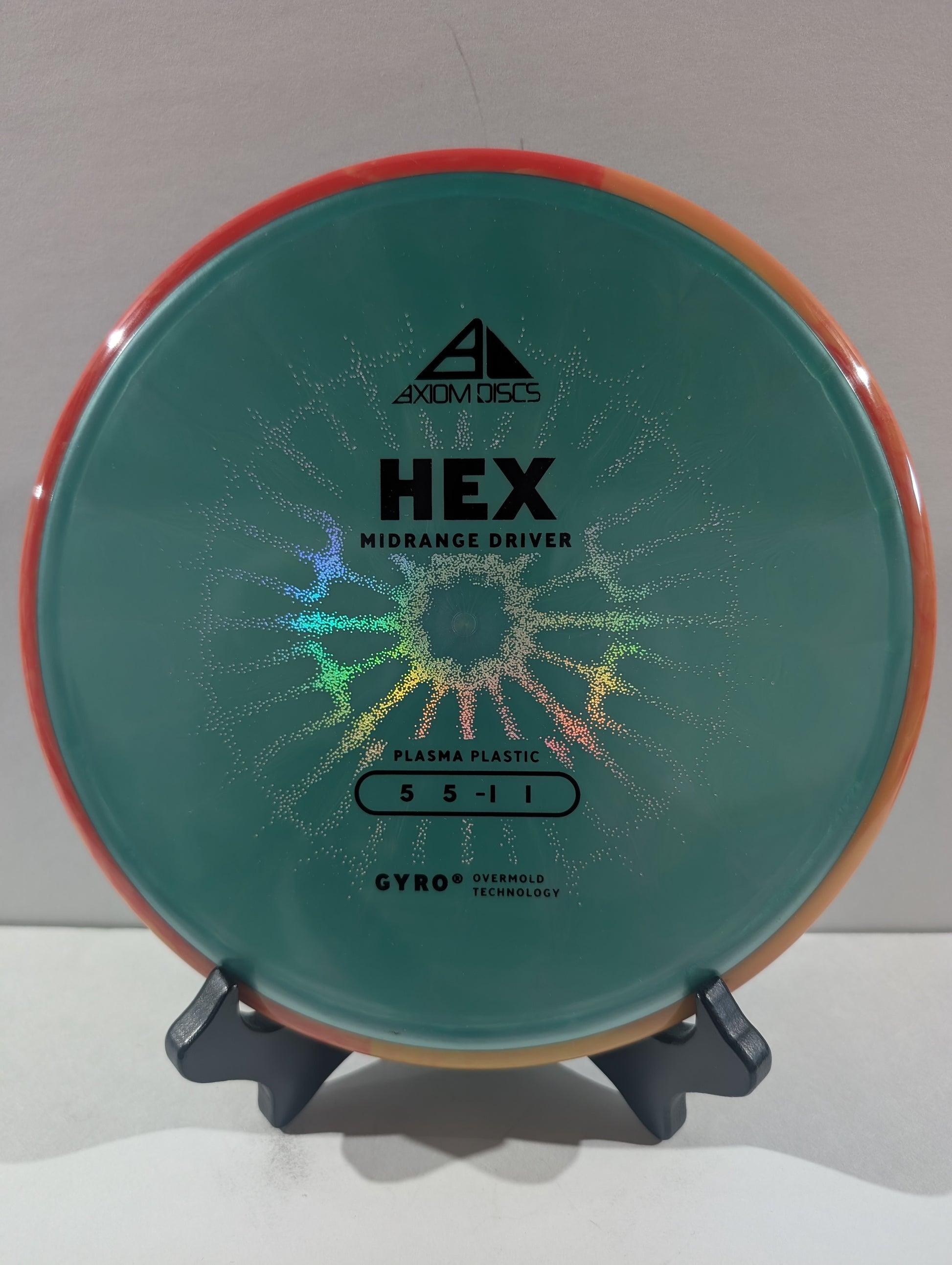 An Axiom Discs Hex Midrange Driver disc golf disc made of Plasma Plastic, featuring a turquoise center with a holographic starburst pattern and an orange rim, displayed on a small stand.