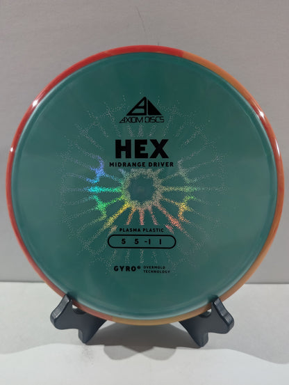 An Axiom Discs Hex Midrange Driver disc golf disc made of Plasma Plastic, featuring a turquoise center with a holographic starburst pattern and an orange rim, displayed on a small stand.