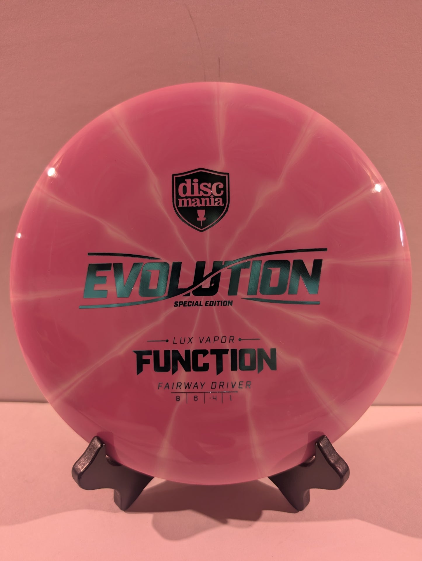 A pink swirled Discmania Evolution Lux Vapor Function fairway driver disc golf disc on a black display stand against a neutral gray background.