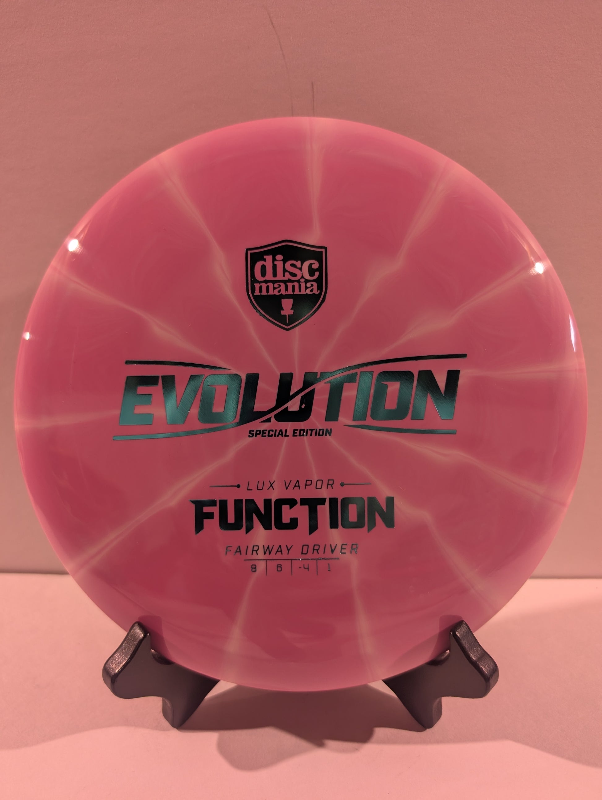 A pink swirled Discmania Evolution Lux Vapor Function fairway driver disc golf disc on a black display stand against a neutral gray background.