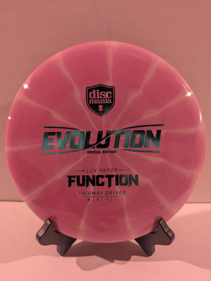 A pink swirled Discmania Evolution Lux Vapor Function fairway driver disc golf disc on a black display stand against a neutral gray background.