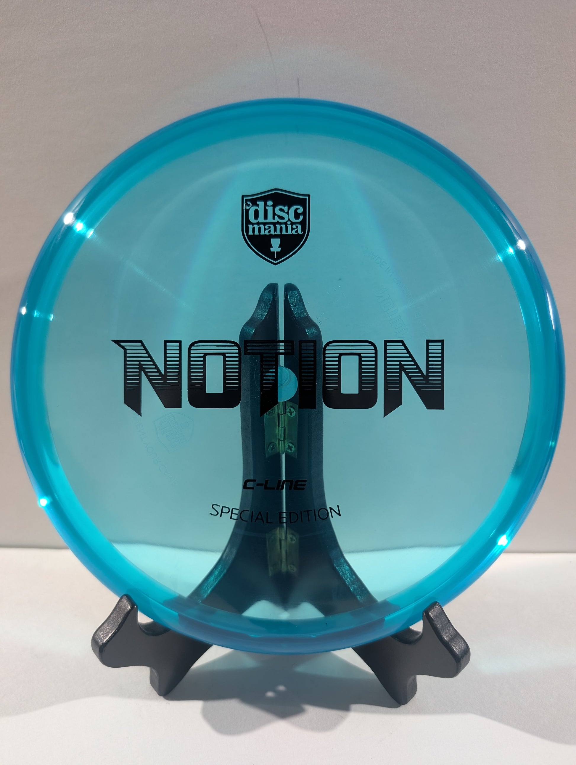 A translucent cyan blue disc golf disc with black printed graphics, displayed on a black stand against a neutral light gray background. The disc features the 'discmania' shield logo and the model name 'NOTION'.