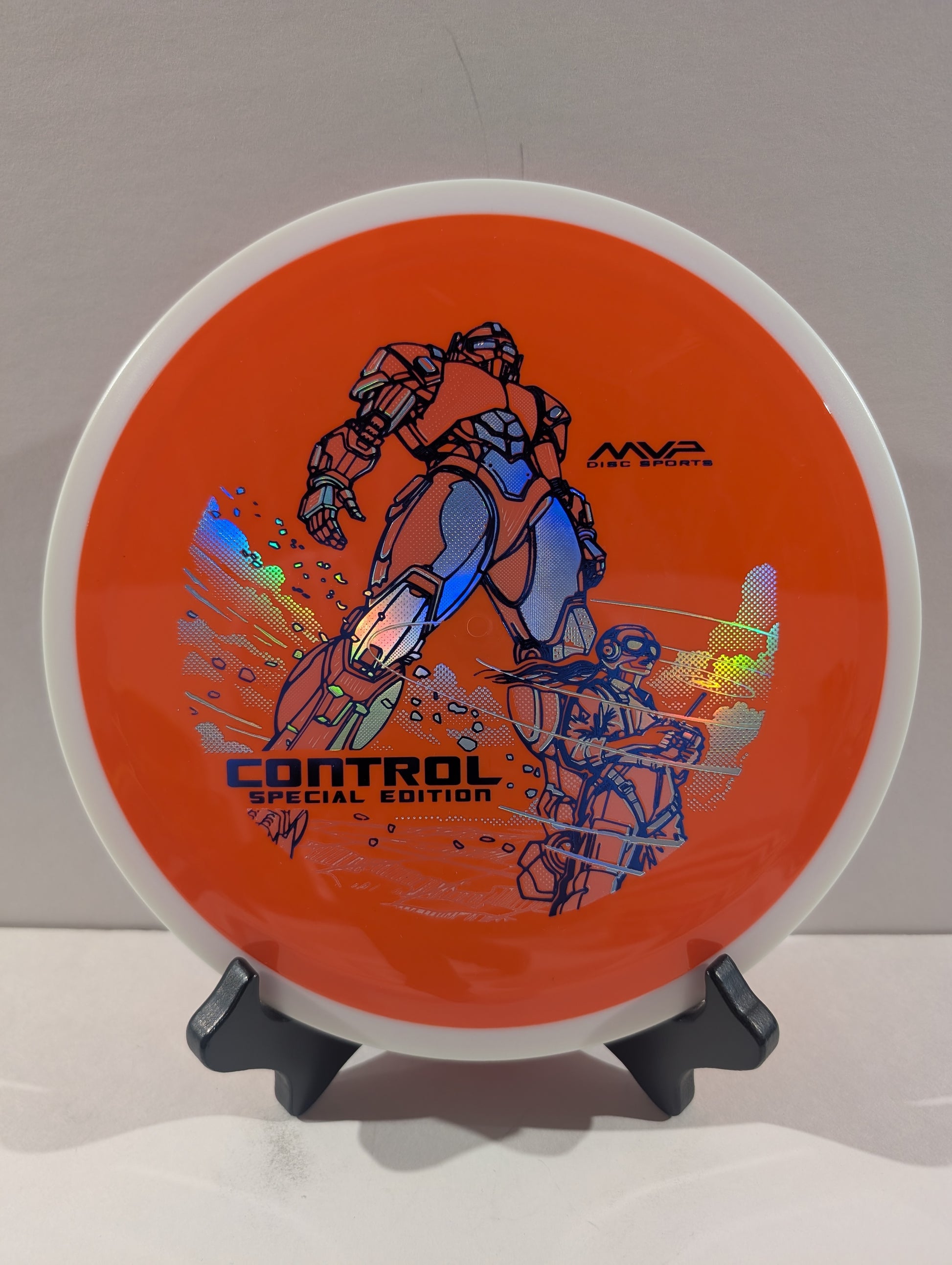 An orange disc golf disc with a white outer rim, featuring a graphic of a robot and the text 'CONTROL SPECIAL EDITION' and 'NWA Disc Sports', displayed on a small black stand.