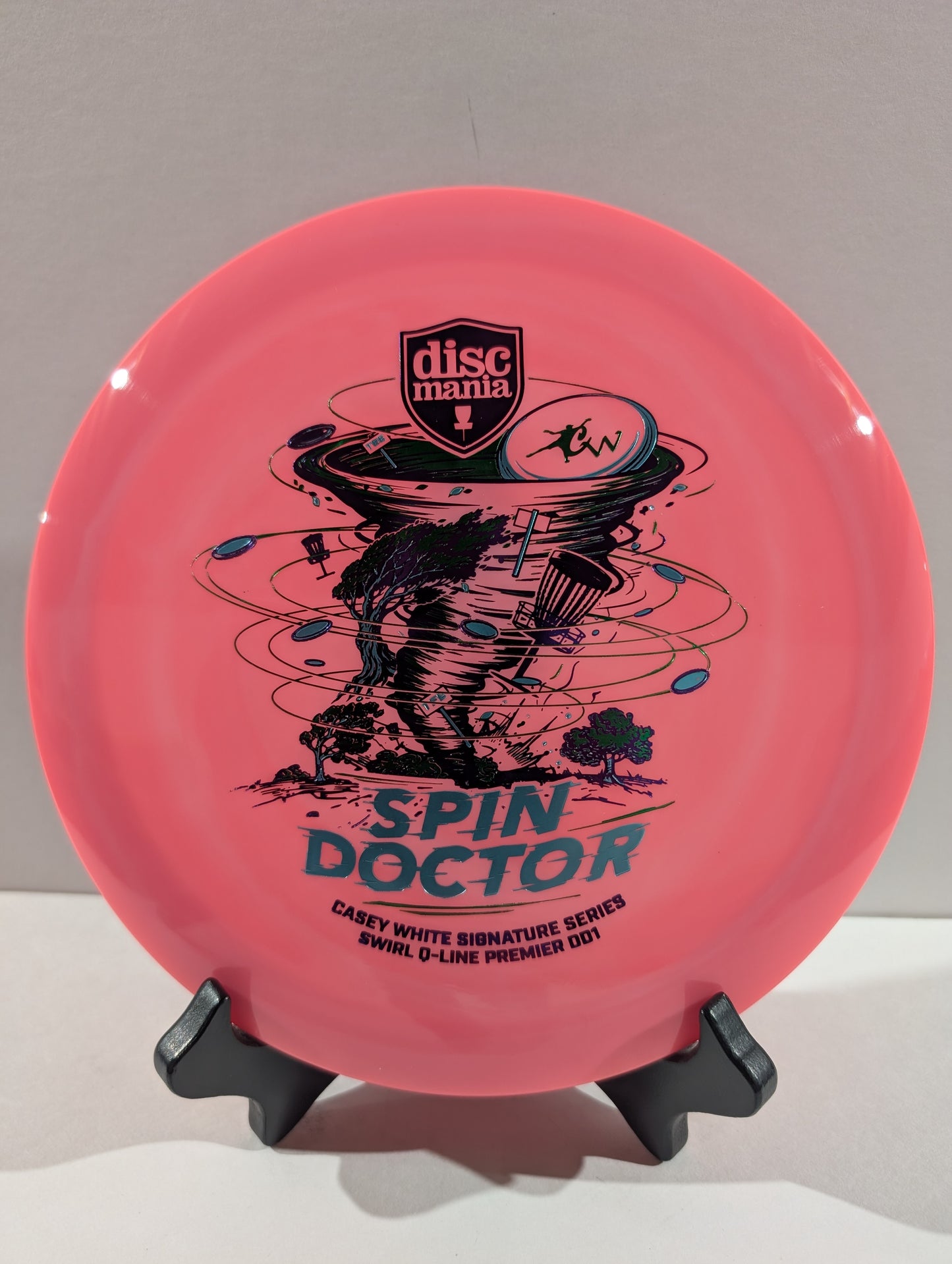 A pink Discmania 'Spin Doctor' disc golf disc displayed on a black stand against a neutral gray background.