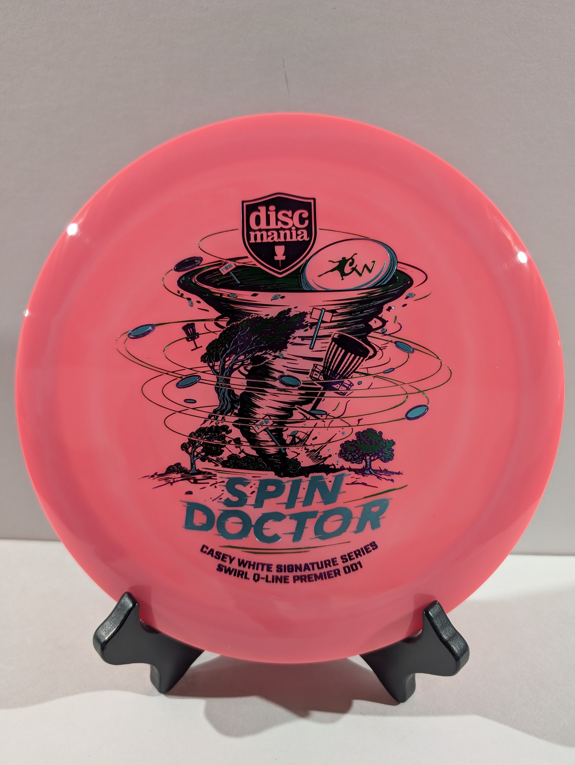 A pink Discmania 'Spin Doctor' disc golf disc displayed on a black stand against a neutral gray background.
