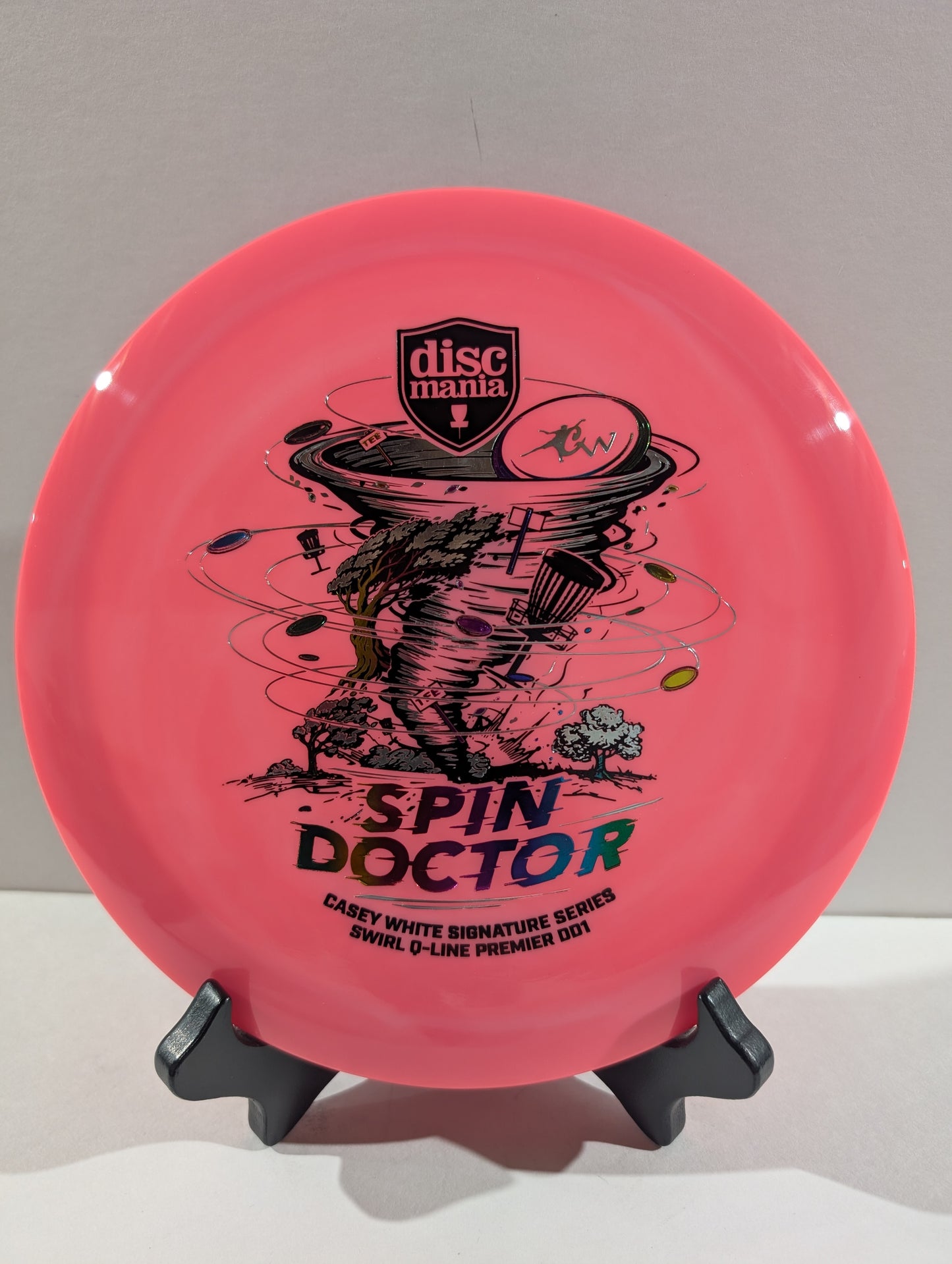 A pink Discmania 'Spin Doctor' disc golf disc displayed on a small black stand against a neutral gray background.