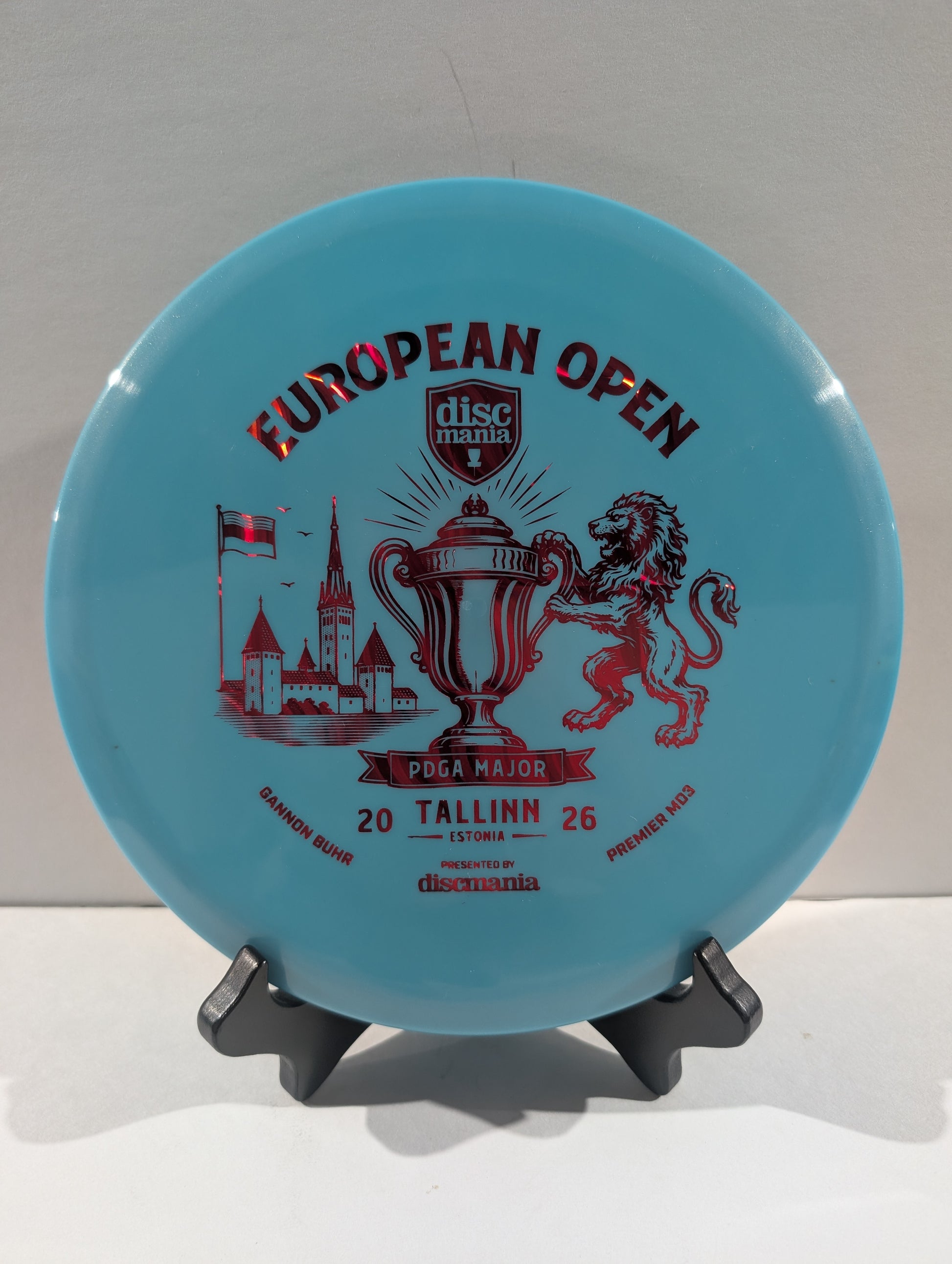 A light blue Discmania European Open disc golf disc displayed on a small black stand against a neutral light gray background.