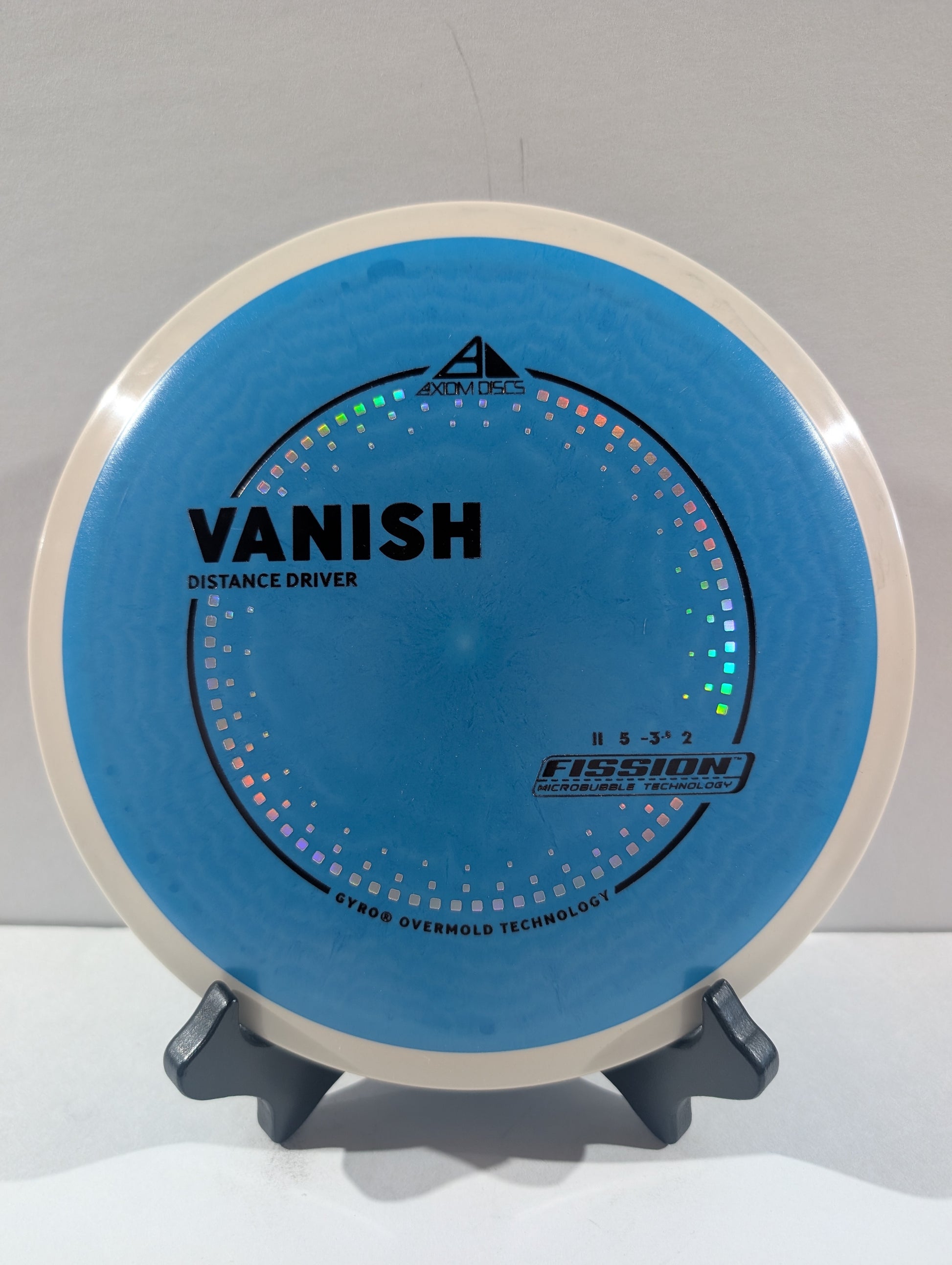 A blue and white disc golf disc, specifically a Vanish distance driver, displayed on a small black stand against a neutral gray background.