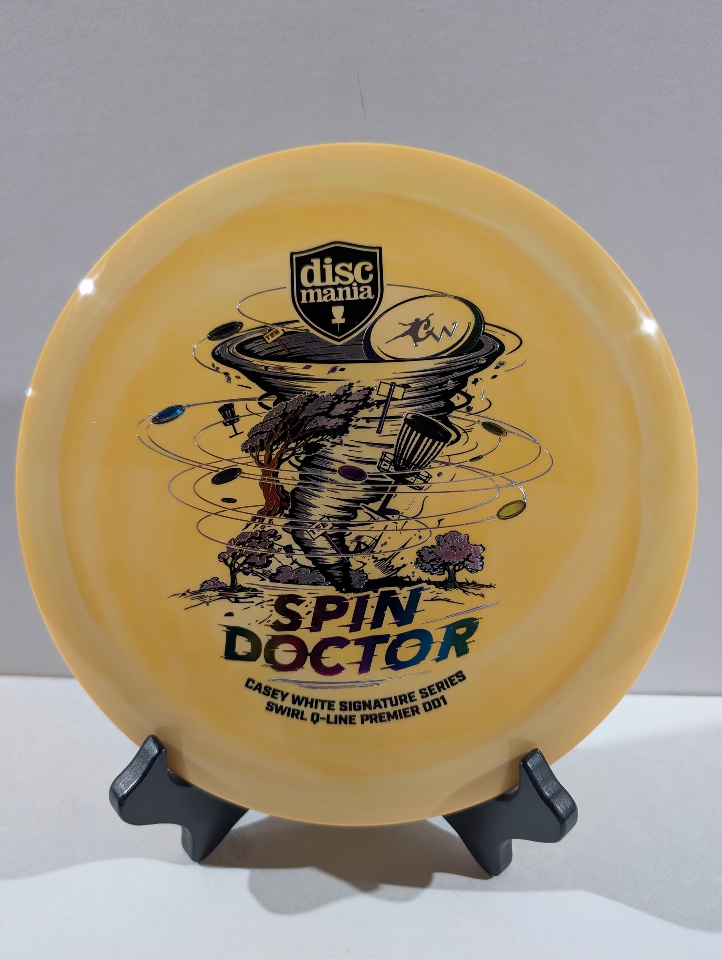 An orange Discmania Spin Doctor disc golf disc displayed on a black stand against a neutral gray background.