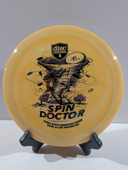 An orange Discmania Spin Doctor disc golf disc displayed on a black stand against a neutral gray background.
