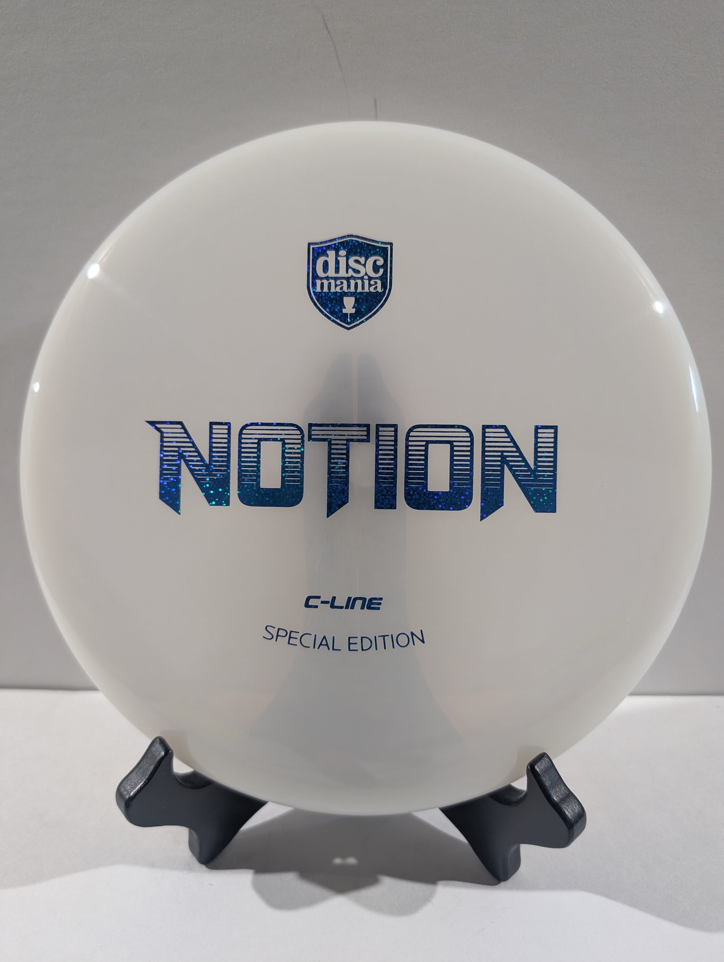 A white Discmania Notion C-Line Special Edition disc golf disc displayed on a black stand against a neutral gray background.