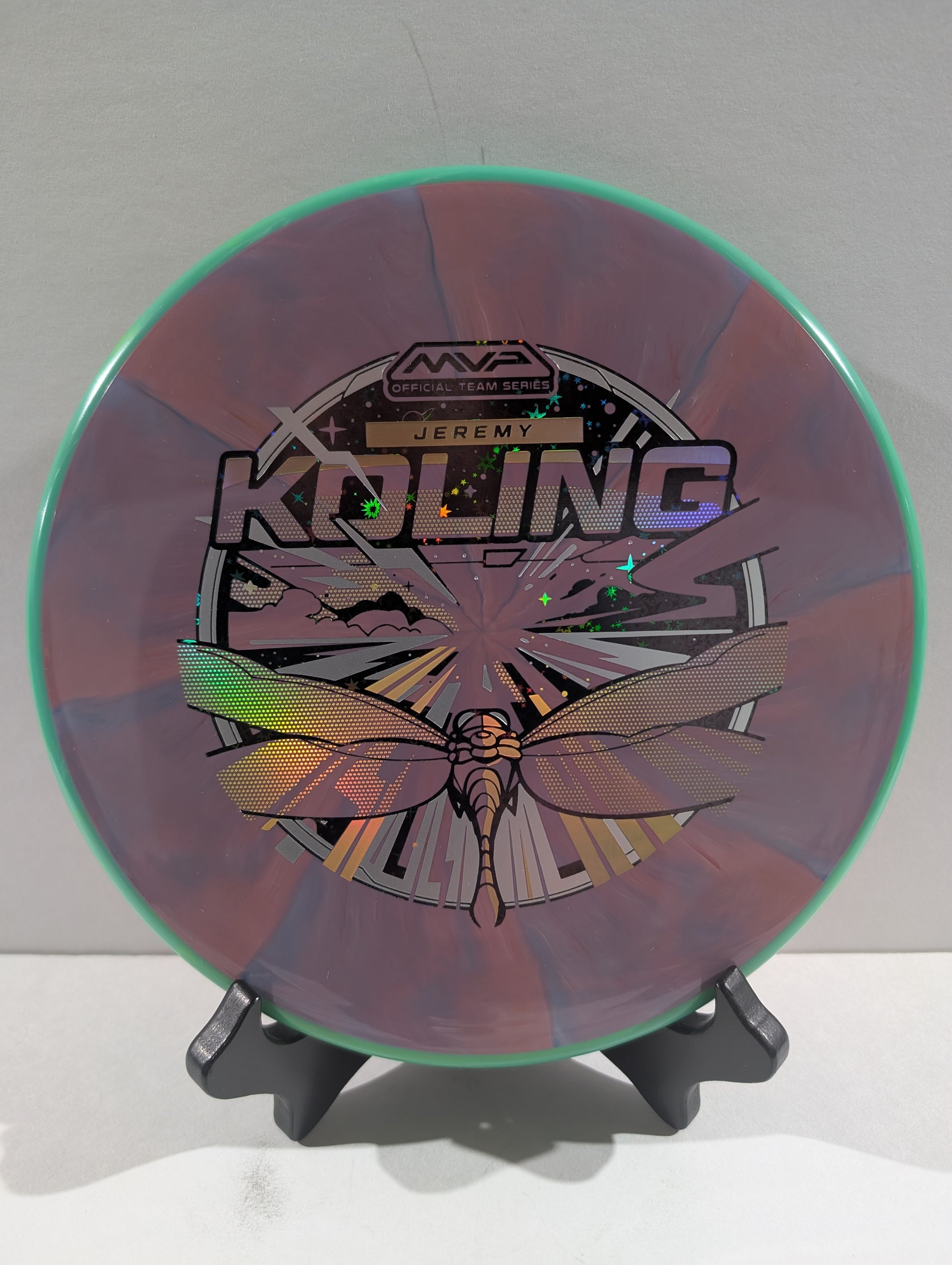 Disc golf disc with a dragonfly design and branding on a stand against a white background