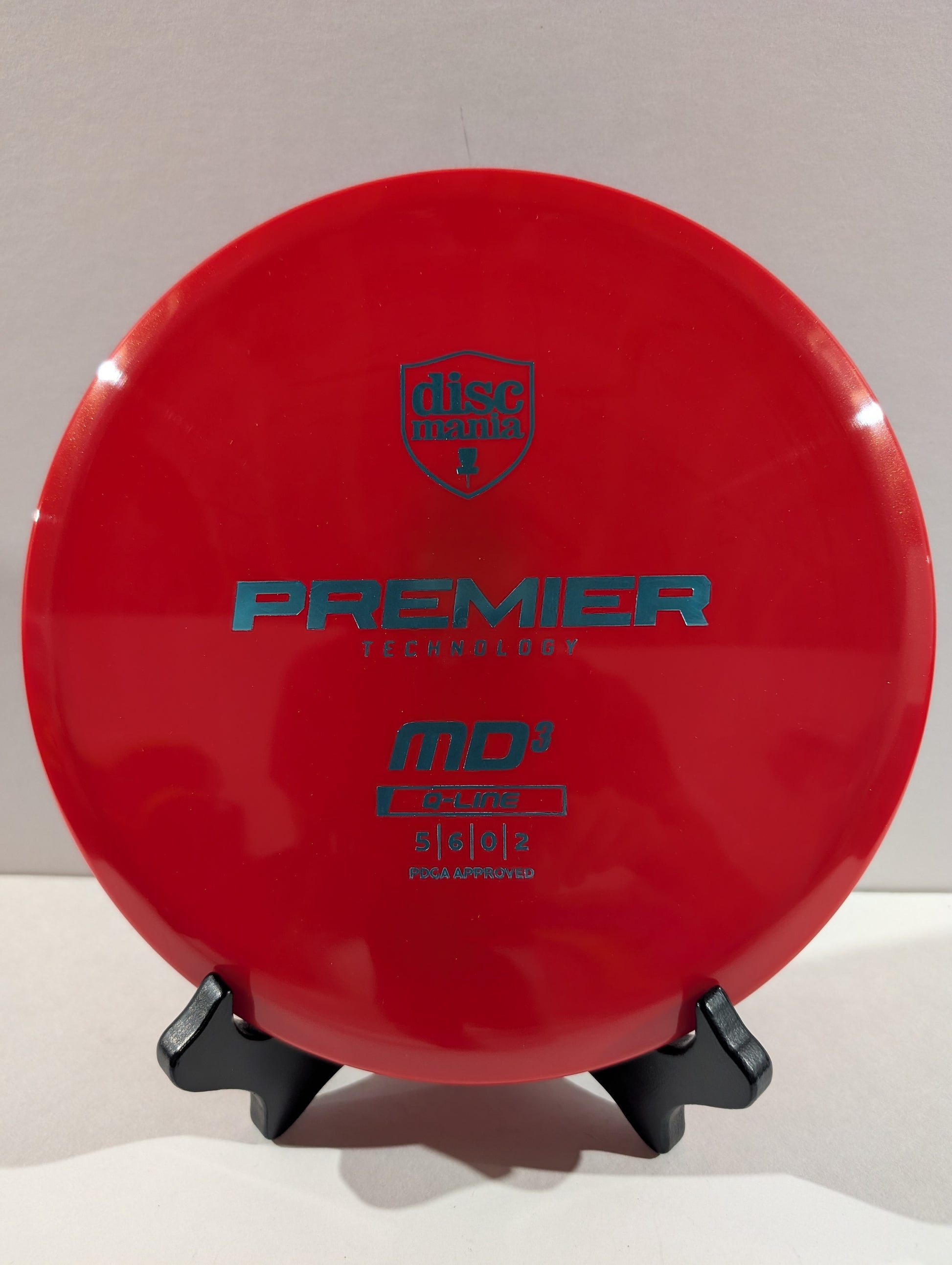 A red Discmania Premier Discraftmer MD3 disc golf disc on a black display stand against a neutral light gray background.