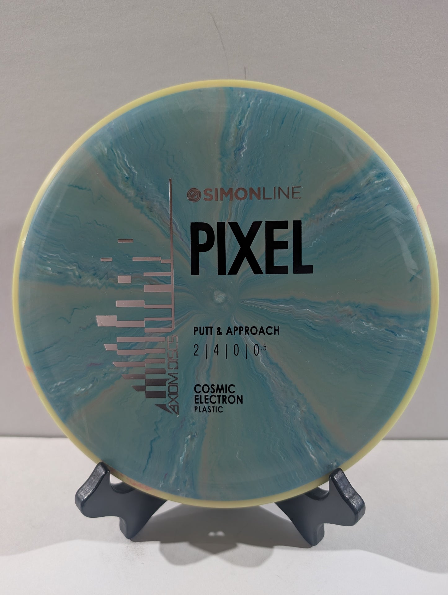 A disc golf disc by Simon Line on a stand, featuring a blue and teal cosmic electron plastic design with a pale yellow outer rim. The disc is labeled 'PIXEL' and 'PUTT & APPROACH'.