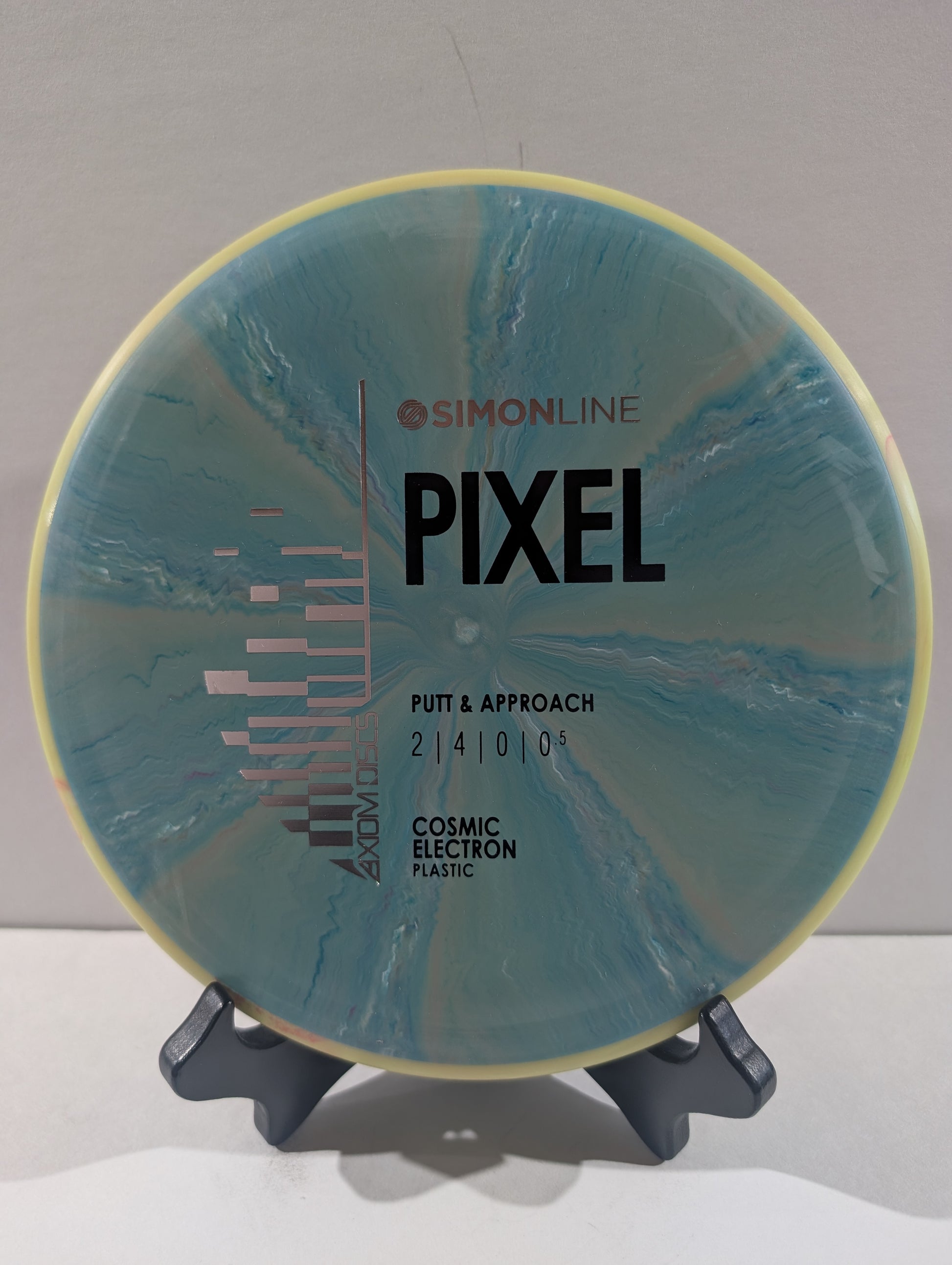 A disc golf disc by Simon Line on a stand, featuring a blue and teal cosmic electron plastic design with a pale yellow outer rim. The disc is labeled 'PIXEL' and 'PUTT & APPROACH'.