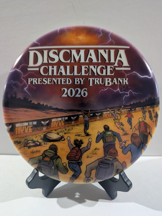 A disc for the Discmania Challenge 2026, presented by TruBank, displayed on a black stand against a neutral light gray background. The disc features a detailed illustration of people at a disc event under a stormy sky, with bold white text at the top.