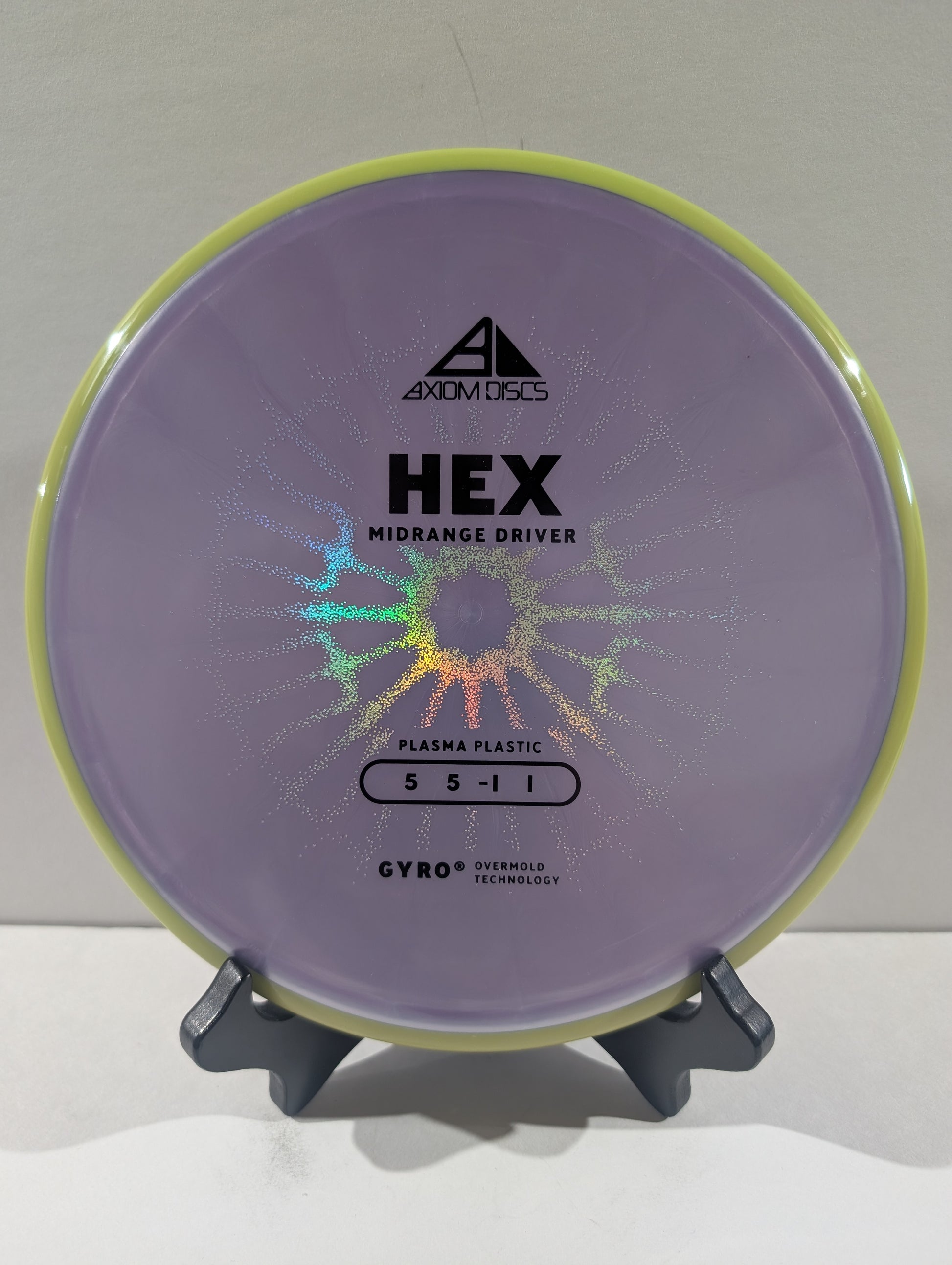 A disc golf disc by Axiom Discs, specifically the HEX Midrange Driver, made of purple Plasma Plastic with a yellow rim, displayed on a small black stand against a neutral light gray background.