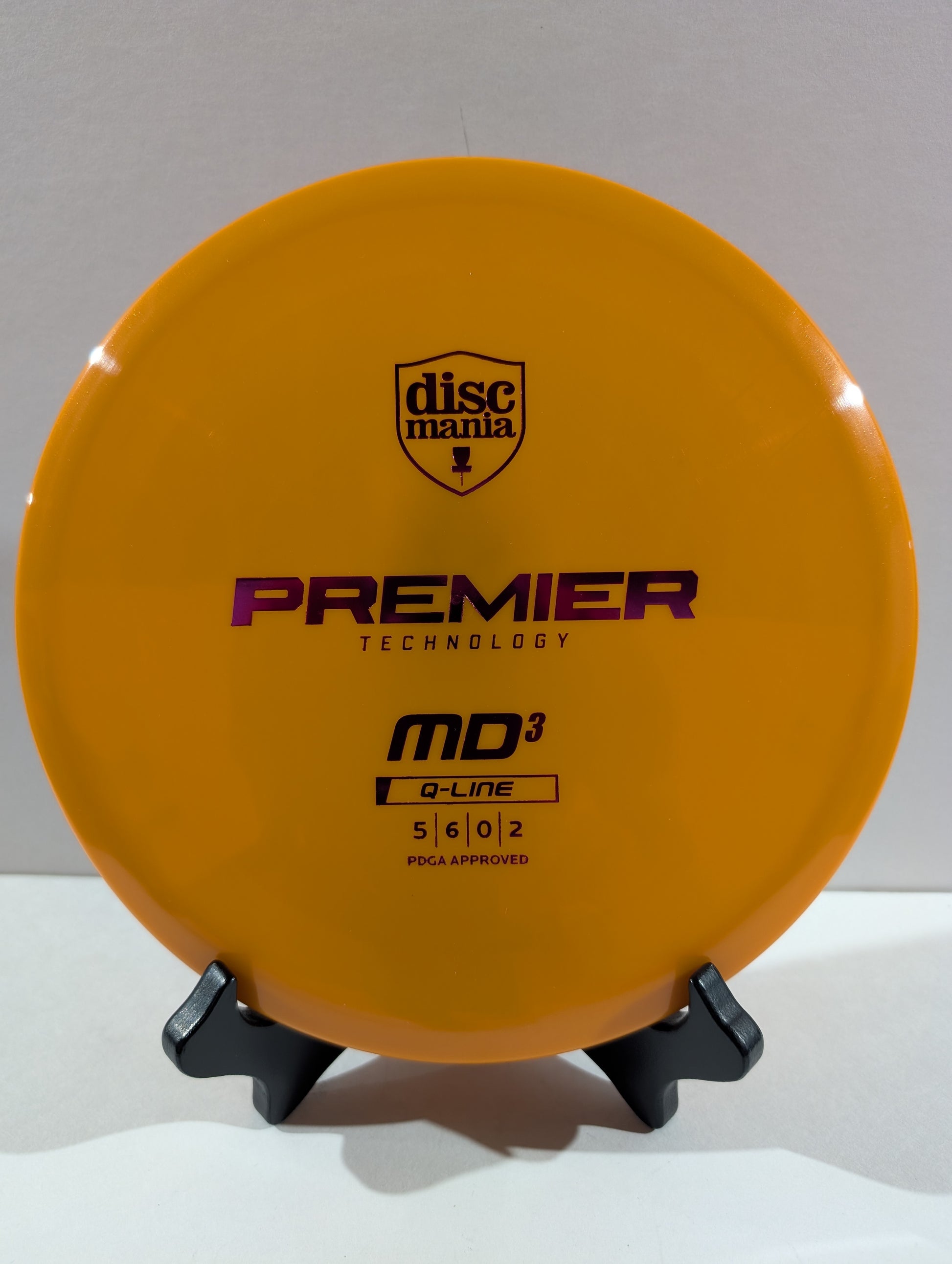 An orange Discmania Premier Technology MD3 Q-Line disc golf disc displayed on a small black stand against a neutral gray background.