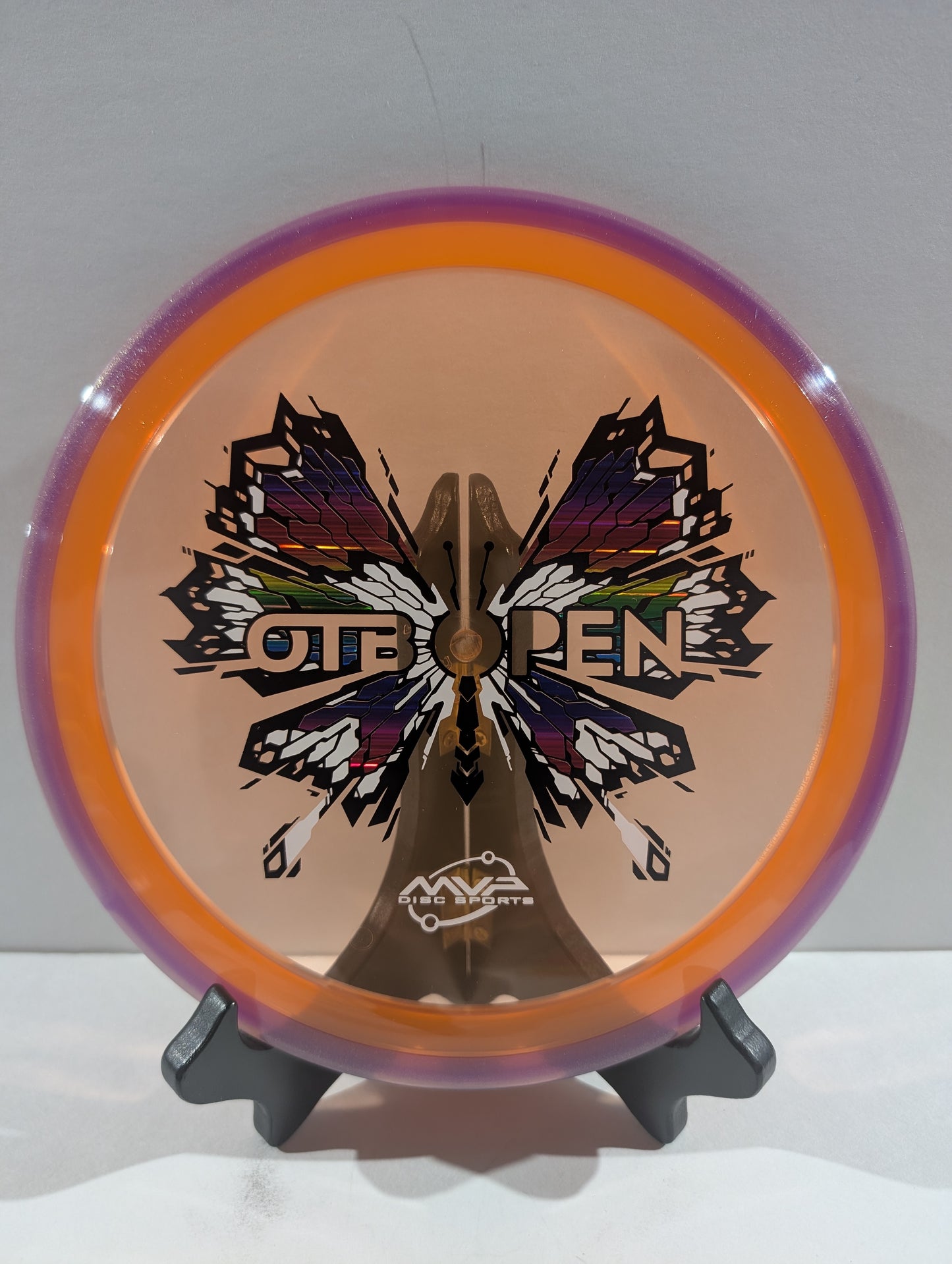 Disc golf disc with butterfly design and 'OTB Open' branding on a stand against a white background